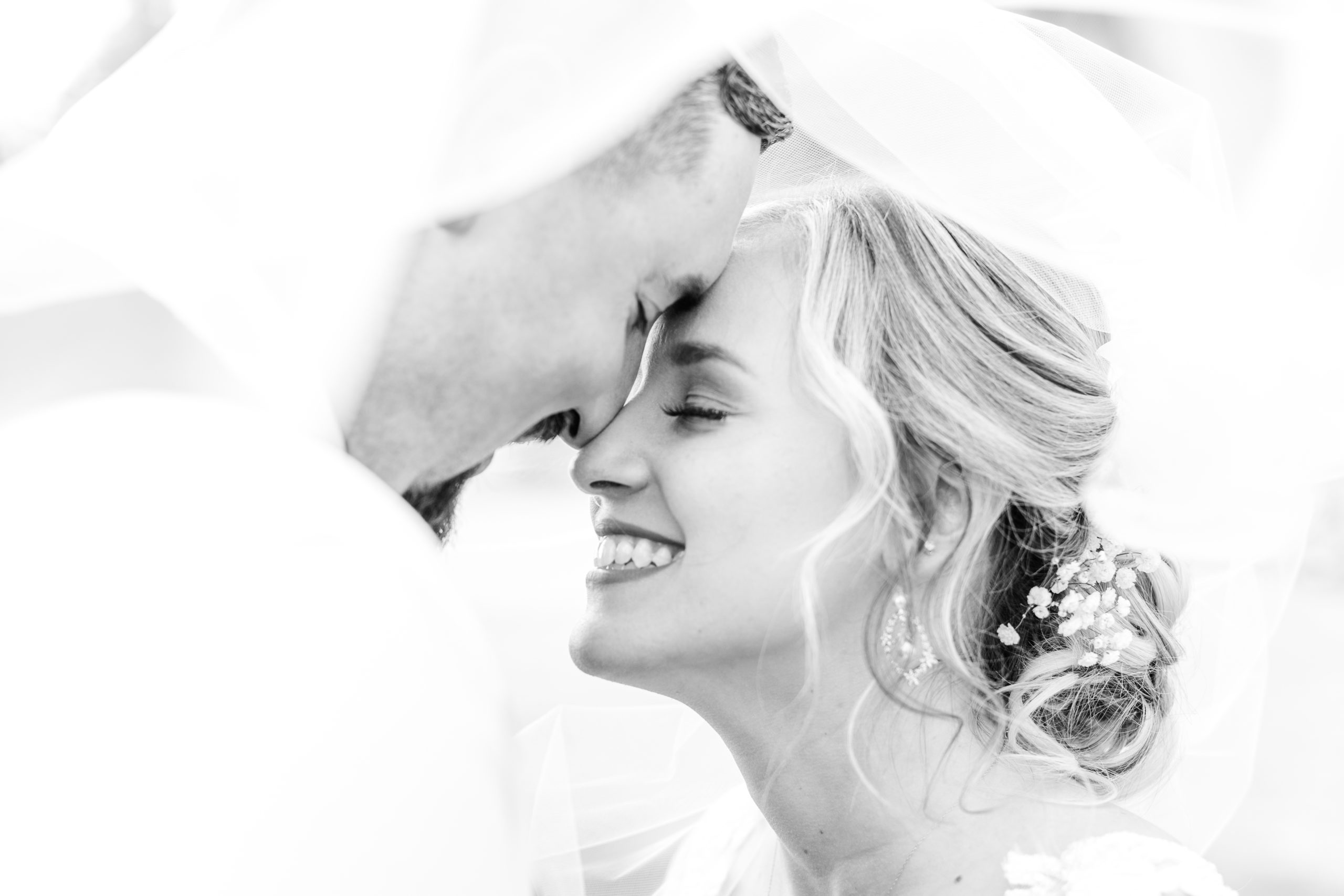 Black and white portrait of bride and groom with foreheads touching and smiling at each other under veil